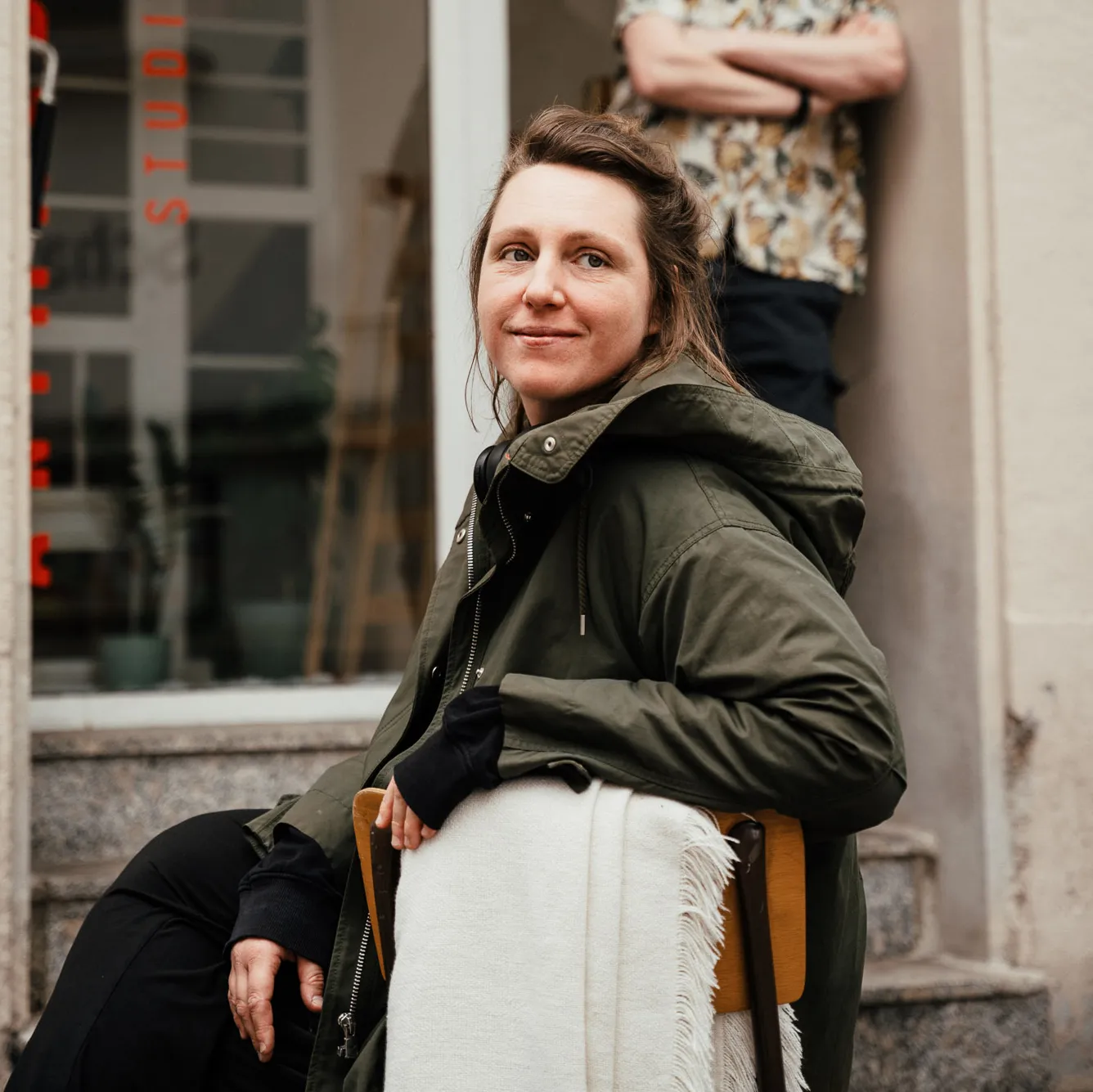 Illustration von Designerin Verena Esterl mit Kopfhörern, blauem Mantel und Schal, umgeben von Pflanzen auf grünem Hintergrund.